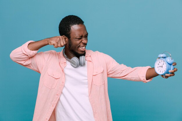 Confiado de pie en pose de lucha sosteniendo reloj despertador joven afroamericano con auriculares en el cuello aislado sobre fondo azul.