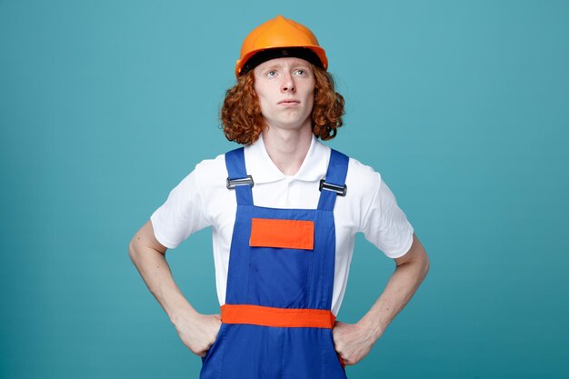 Confiado poniendo las manos en las caderas joven constructor hombre en uniforme aislado sobre fondo azul.