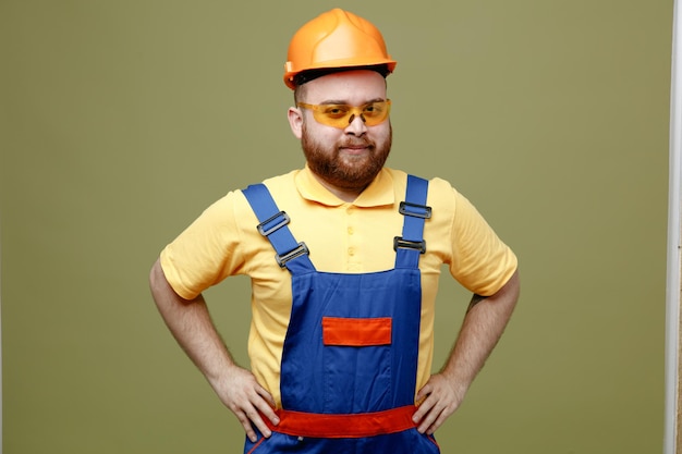 Confiado poniendo las manos en las caderas joven constructor hombre en uniforme aislado sobre fondo verde