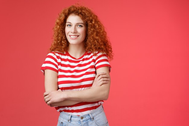 Confiado tierna pelirroja tonta hermosa chica de pelo rizado con brazos cruzados cuerpo sonriendo ampliamente encantada divirtiéndose alegremente disfrutar de la cálida noche de verano camina sobre fondo rojo de vacaciones en el campo tropical.