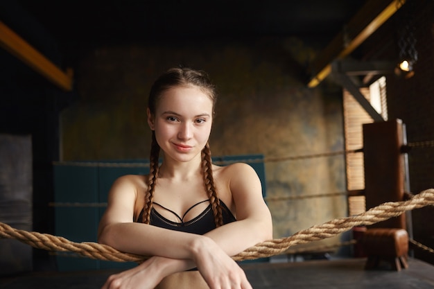 Confianza adolescente con dos trenzas de pie en el ring de boxeo, esperando a su instructor o entrenador, sonriendo a la cámara, cruzando los brazos en una cuerda frente a ella