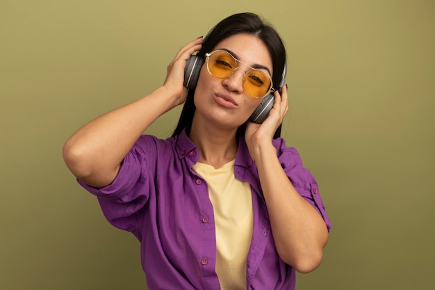 Confianza bastante morena chica caucásica en gafas de sol con auriculares mira a la cámara en verde oliva