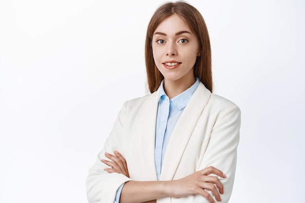 Confianza empresaria joven en traje, brazos cruzados sobre el pecho y sonrisa al frente, mirada determinada y hábil, de pie sobre la pared blanca