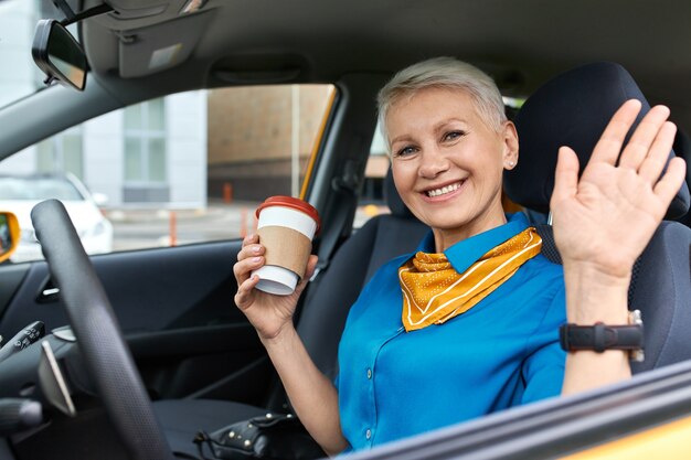 Confianza empresaria madura alegre con cabello rubio corto sentado en el fiador del conductor sosteniendo un vaso de papel desechable