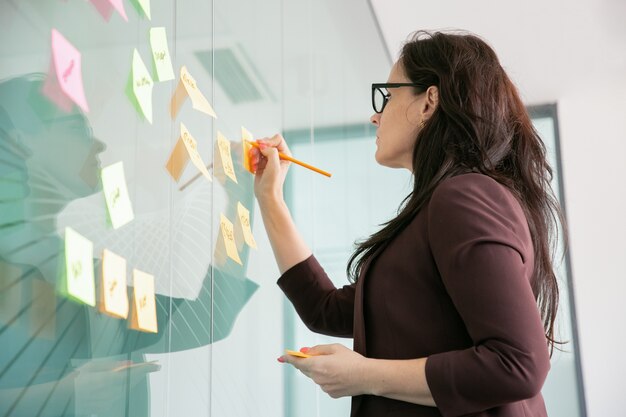 Confianza empresaria de mediana edad escribiendo en la etiqueta engomada con lápiz y lluvia de ideas