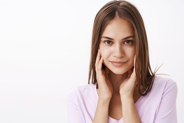 Confianza femenina atractiva morena femenina con cabello lacio y piel limpia tocando la cara suavemente y sonriente coqueteando atrevido sobre la pared blanca