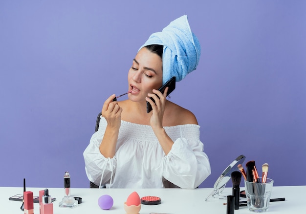 Confianza hermosa chica envuelta toalla para el cabello se sienta en la mesa con herramientas de maquillaje sosteniendo y aplicando brillo de labios hablando por teléfono aislado en la pared púrpura