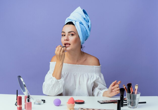 Confianza hermosa chica envuelta toalla para el cabello se sienta en la mesa con herramientas de maquillaje sosteniendo y aplicando lápiz labial mirando aislado en la pared púrpura