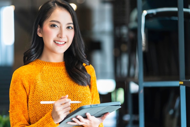 Confianza inteligente, empresaria asiática, empresaria nueva, propietaria de una pequeña empresa, mujer de negocios, ropa informal inteligente, sonrisa, uso de la mano, tableta, comprobación de inventario en la oficina de la sala de exposición, fondo diurno