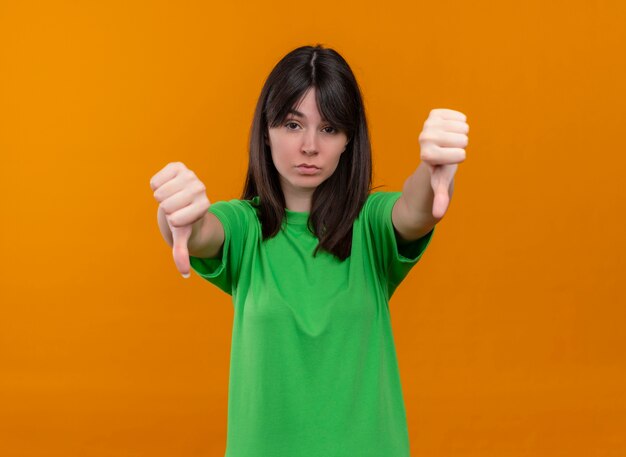 Confianza joven caucásica en camisa verde pulgares hacia abajo con ambas manos sobre fondo naranja aislado