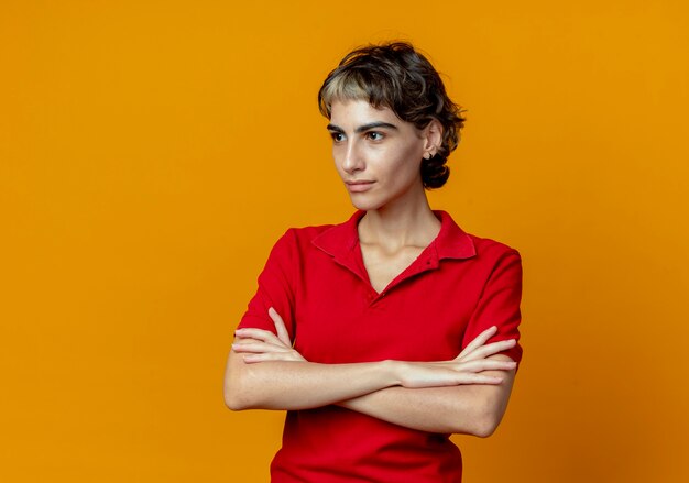 Confianza joven caucásica con corte de pelo pixie de pie con postura cerrada mirando al lado aislado sobre fondo naranja con espacio de copia