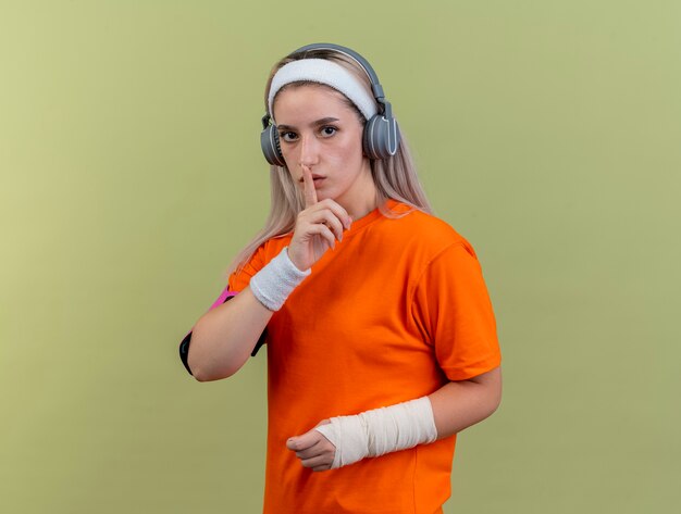 Confianza joven caucásica deportiva con tirantes en los auriculares con muñequeras de diadema y gestos de brazalete de teléfono signo de silencio