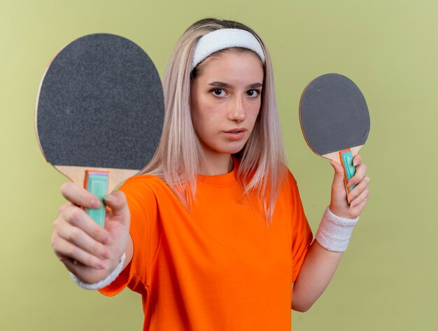 Confianza joven caucásica deportiva con tirantes con diadema y muñequeras sosteniendo raquetas de ping-pong