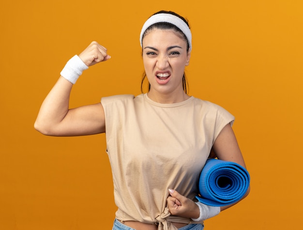 Confianza joven caucásica deportiva vistiendo diadema y muñequeras sosteniendo estera de fitness mirando al frente haciendo un gesto fuerte aislado en la pared naranja