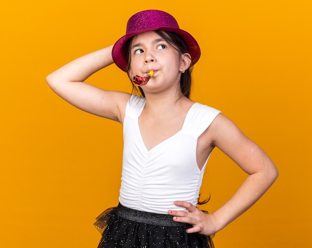 Confianza joven caucásica con gorro de fiesta púrpura poniendo la mano en la cabeza y soplando el silbato del partido mirando al lado aislado en la pared naranja con espacio de copia