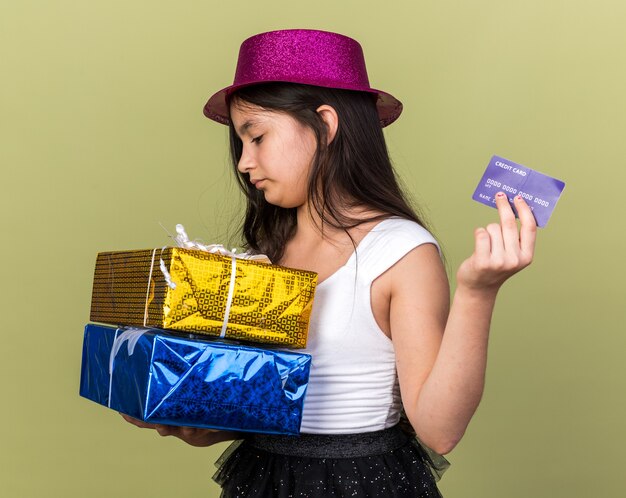 Confianza joven caucásica con gorro de fiesta púrpura sosteniendo una tarjeta de crédito y mirando cajas de regalo aisladas en la pared verde oliva con espacio de copia