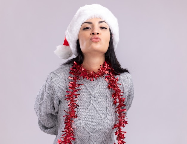 Foto gratuita confianza joven caucásica con sombrero de navidad y guirnalda de oropel alrededor del cuello manteniendo las manos detrás de la espalda mirando a la cámara haciendo gesto de beso aislado sobre fondo blanco con espacio de copia