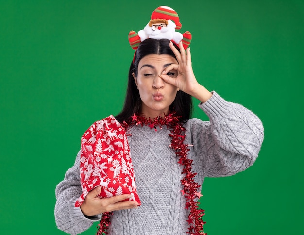 Confianza joven caucásica vistiendo santa claus diadema y guirnalda de oropel alrededor del cuello sosteniendo un saco de regalo de Navidad haciendo gesto de mirada frunciendo los labios con un ojo cerrado aislado en la pared verde