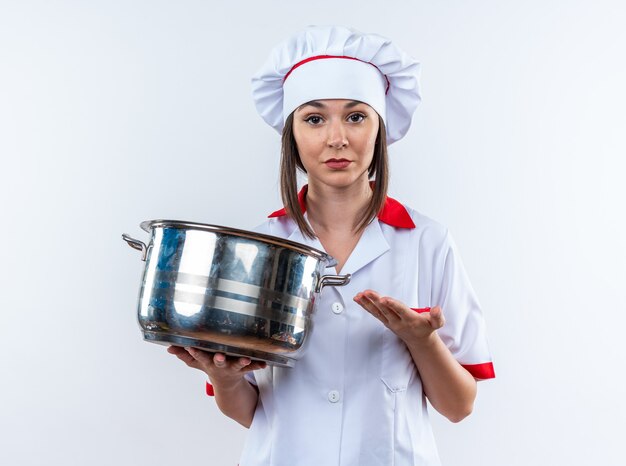 Confianza joven cocinera vistiendo uniforme de chef sosteniendo y puntos en una cacerola aislado en la pared blanca