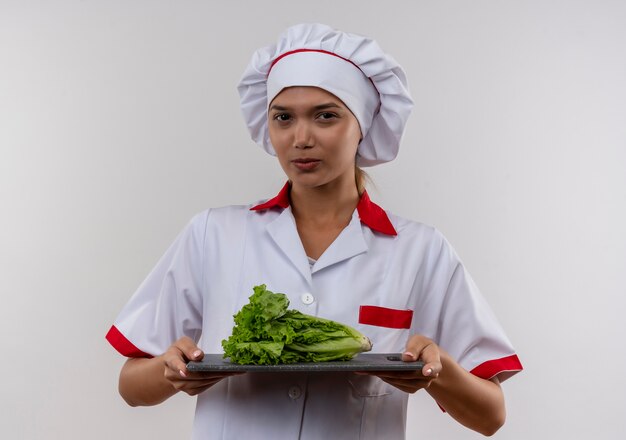 Confianza joven cocinera vistiendo uniforme de chef sosteniendo sald sobre tabla de cortar en pared blanca aislada