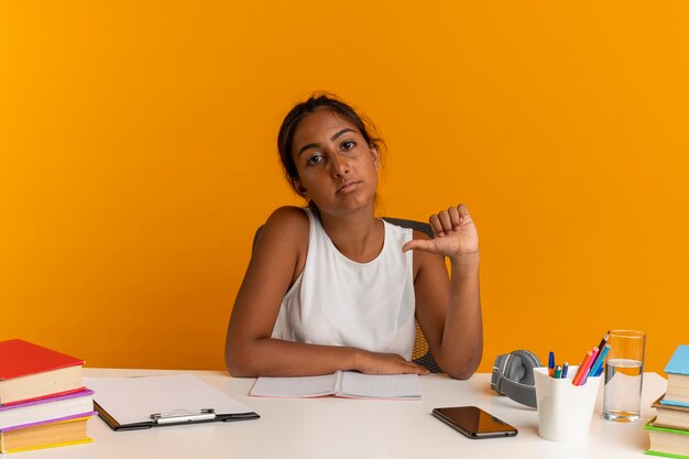 Confianza joven colegiala sentada en un escritorio con herramientas escolares apunta a sí misma