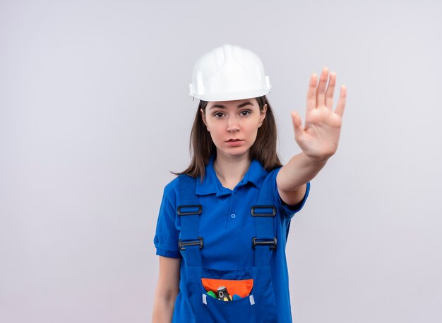 Confianza joven constructor con casco de seguridad blanco y gestos uniformes azules se detienen sobre fondo blanco aislado con espacio de copia