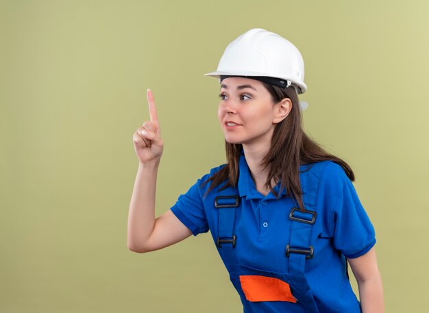 Confianza joven constructor con casco de seguridad blanco y uniforme azul apunta hacia arriba sobre fondo verde aislado con espacio de copia