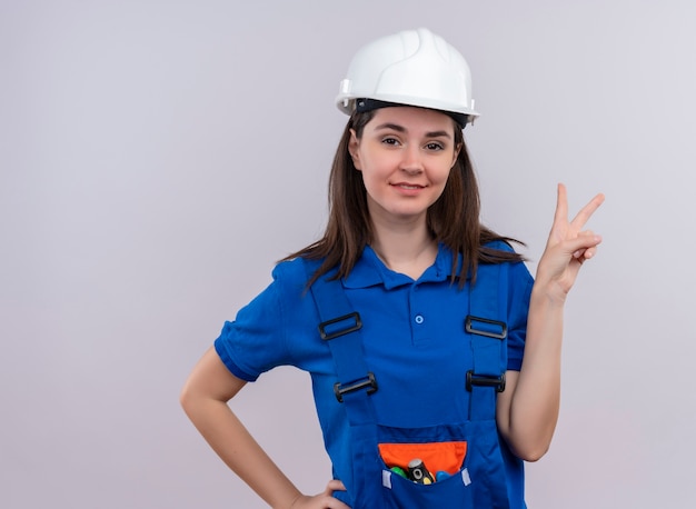 Confianza joven constructor con casco de seguridad blanco y uniforme azul hace gestos de signo de victoria y pone la mano en la cintura sobre fondo blanco aislado con espacio de copia