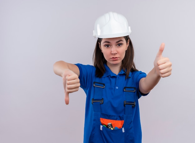 Foto gratuita confianza joven constructor con casco de seguridad blanco y uniforme azul pulgar hacia abajo y pulgar hacia arriba sobre fondo blanco aislado con espacio de copia