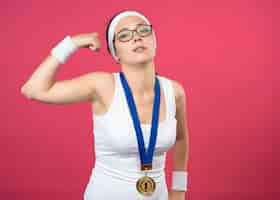 Foto gratuita confianza joven deportiva en gafas ópticas con medalla de oro alrededor del cuello con diadema y muñequeras tensa bíceps