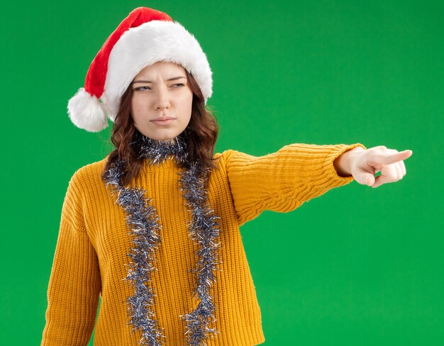 Confianza joven eslava con gorro de Papá Noel y con guirnalda alrededor del cuello mirando y apuntando al lado aislado sobre fondo verde con espacio de copia