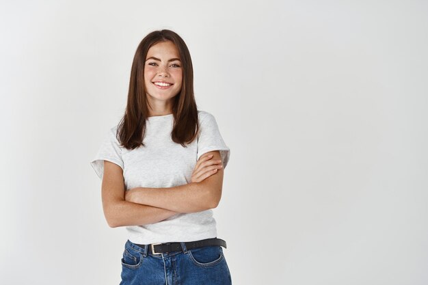 Confianza joven estudiante de pie con los brazos cruzados y sonriendo al frente, posando sobre la pared blanca