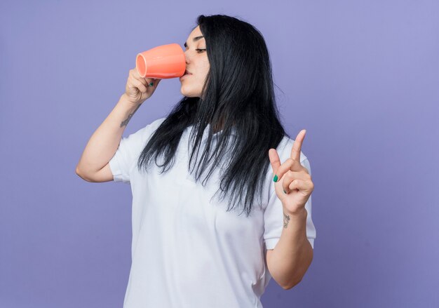 Confianza joven morena caucásica bebe de la taza apuntando hacia arriba aislado en la pared púrpura