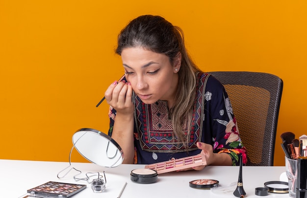 Confianza joven morena sentada en la mesa con herramientas de maquillaje aplicando sombra de ojos con pincel de maquillaje mirando en el espejo