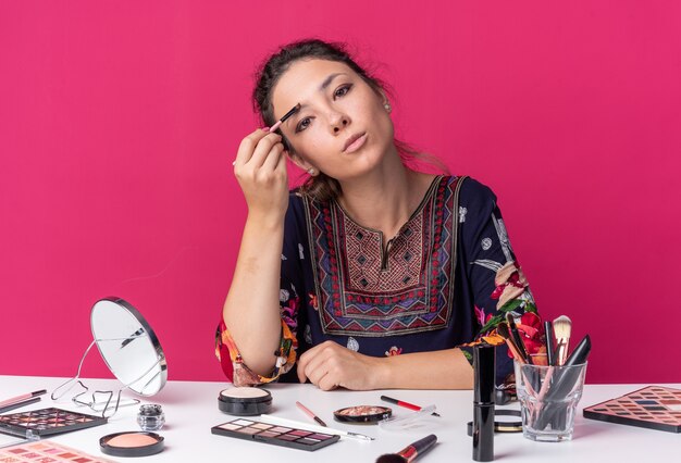 Confianza joven morena sentada a la mesa con herramientas de maquillaje aplicando sombra de ojos con pincel de maquillaje