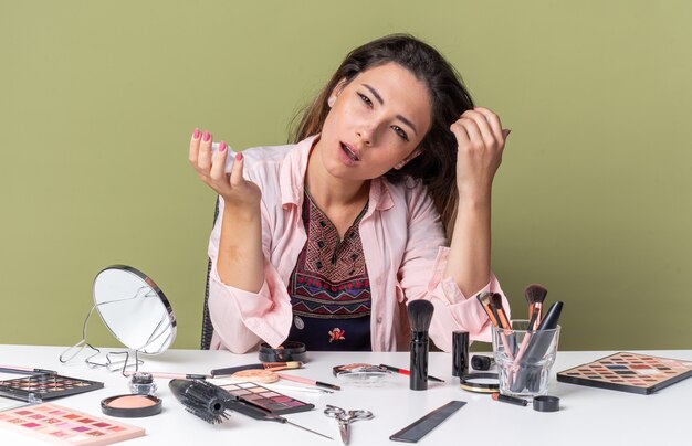 Confianza joven morena sentada a la mesa con herramientas de maquillaje sosteniendo mousse para el cabello aislado en la pared verde oliva con espacio de copia