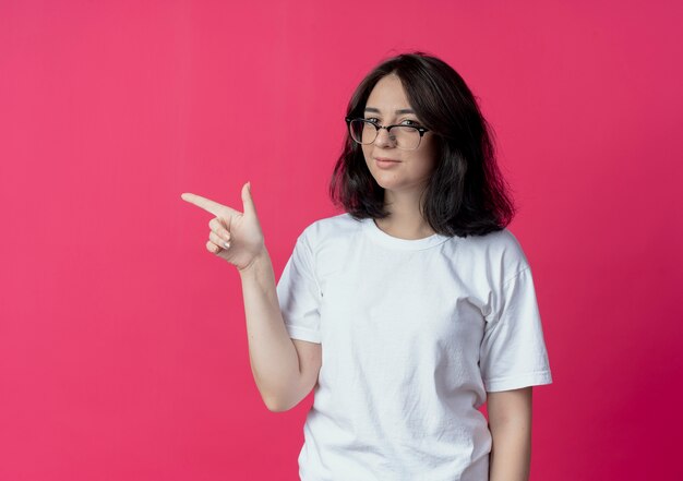 Confianza joven mujer bonita con gafas apuntando al lado de la pared carmesí