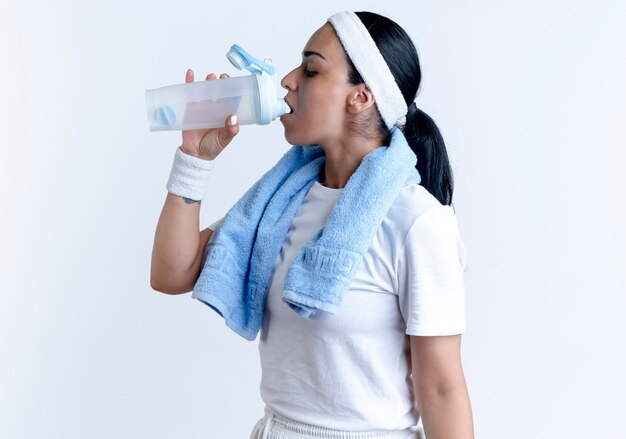 Confianza joven mujer deportiva caucásica vistiendo muñequeras diadema con una toalla en el cuello bebe agua