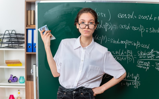 Foto gratuita confianza joven profesora de matemáticas con gafas de pie delante de la pizarra manteniendo la mano en la cintura sosteniendo el borrador de la pizarra mirando al frente en el aula