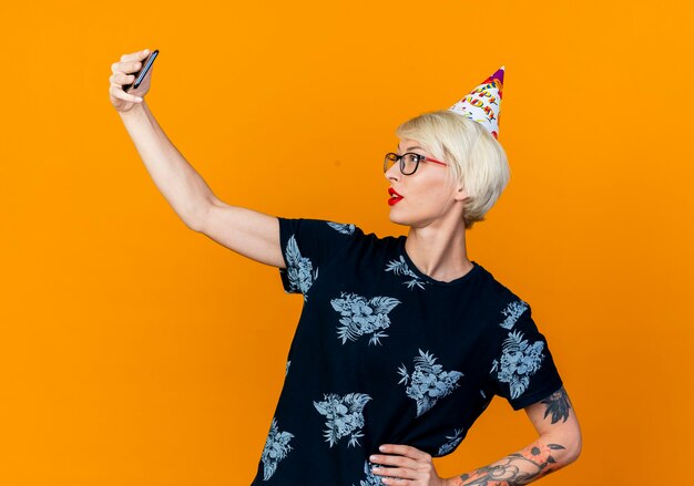 Confianza joven rubia fiestera con gafas y gorro de cumpleaños tomando selfie manteniendo la mano en la cintura aislada sobre fondo naranja