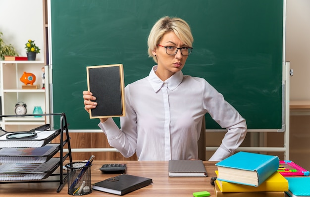 Confianza joven rubia profesora con gafas sentado en el escritorio con herramientas escolares en el aula mostrando mini pizarra manteniendo la mano en la cintura mirando a la cámara