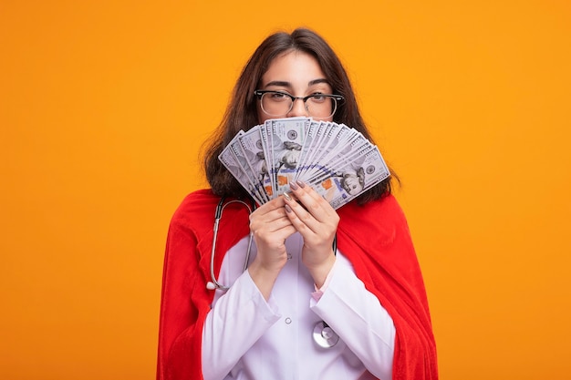 Confianza joven superhéroe caucásica vestida con uniforme médico y un estetoscopio con gafas sosteniendo dinero detrás de él aislado en la pared con espacio de copia