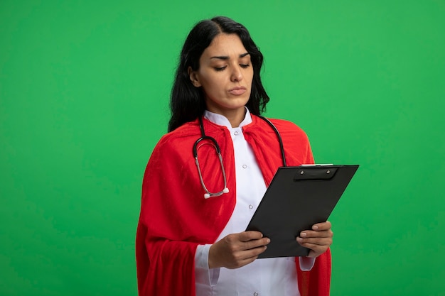 Confianza joven superhéroe vestida con bata médica con estetoscopio sosteniendo y mirando el portapapeles aislado en verde