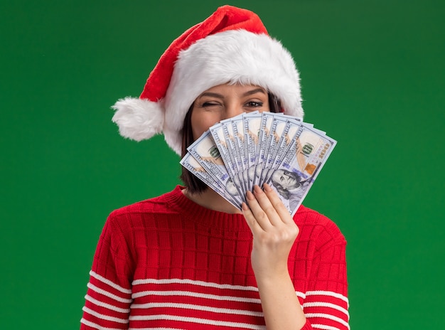 Confianza joven vistiendo gorro de Papá Noel con dinero detrás de él guiñando un ojo aislado en la pared verde