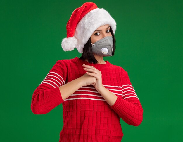 Confianza joven vistiendo gorro de Papá Noel y máscara protectora mirando a la cámara haciendo gesto de saludo aislado sobre fondo verde