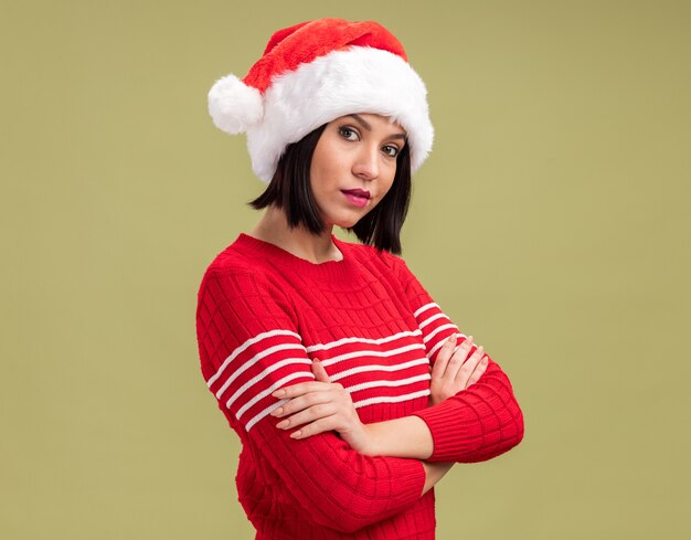 Confianza joven vistiendo gorro de Papá Noel de pie en la vista de perfil mirando a la cámara aislada sobre fondo verde oliva con espacio de copia