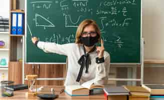 Foto gratuita confianza maestra joven con gafas y máscara médica se sienta a la mesa con herramientas escolares puntos en la pizarra con puntero en el aula