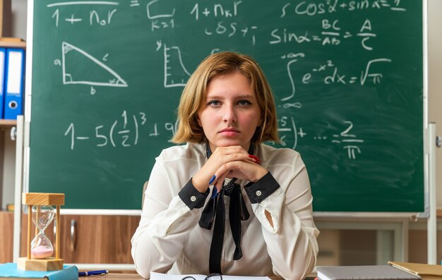 Confianza mirando al frente joven maestra se sienta a la mesa con útiles escolares en el aula