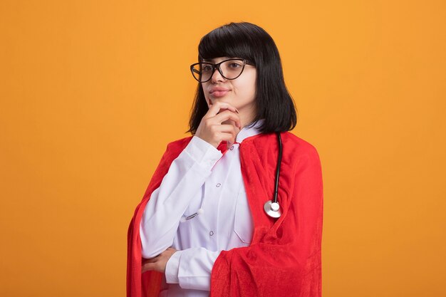 Confianza mirando al lado joven superhéroe chica con estetoscopio con bata médica y manto con gafas agarró la barbilla