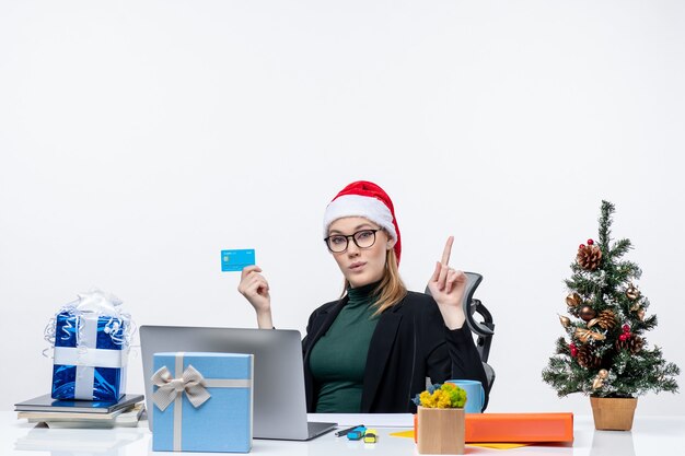 Confianza mujer atractiva ambiciosa con sombrero de santa claus y gafas sentado en una mesa y sosteniendo una tarjeta bancaria y apuntando arriba en la oficina